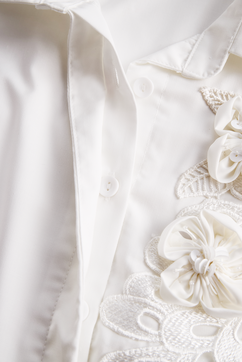 FJØRNA white sleeveless women’s blouse with floral embroidery and 3D appliqué detailing, shown on a plain background.