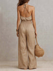 FJØRNA tan cotton linen sleeveless jumpsuit with halter neck, smocked waist, and wide-leg design, perfect for summer wear.