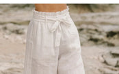 FJØRNA linen wide-leg women's trousers with elastic waist and ruffle detail, shown on a model in a bright, airy setting.