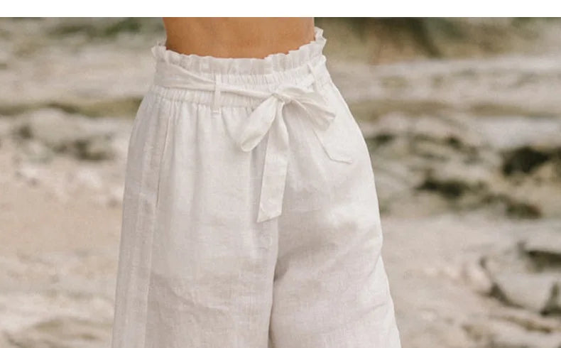 FJØRNA linen wide-leg women's trousers with elastic waist and ruffle detail, shown on a model in a bright, airy setting.