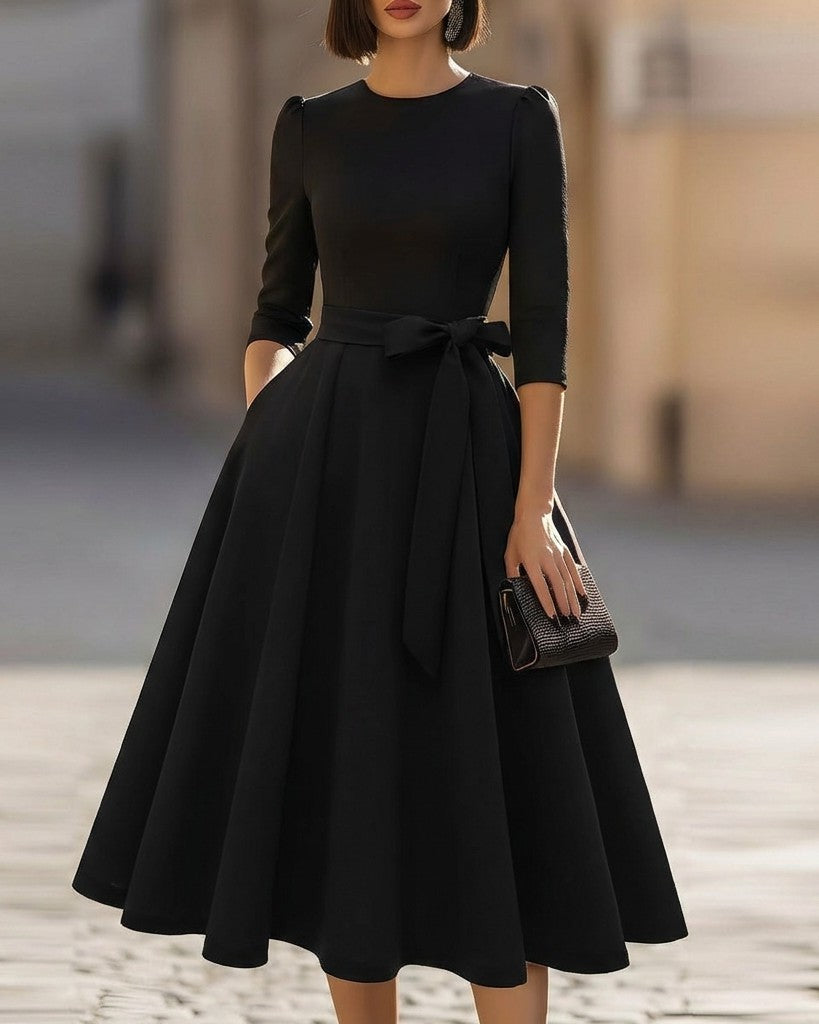 FJØRNA crew-neck A-line midi dress, elegant wedding guest dress in soft fabric, shown on a model with floral background.