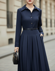 FJØRNA collared button-front midi dress with tailored pleats and belted waist, shown on a model in a stylish indoor setting.