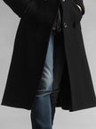 Black FJØRNA midi wool coat with pockets for women, stylish and warm, shown hanging on a wooden hanger.
